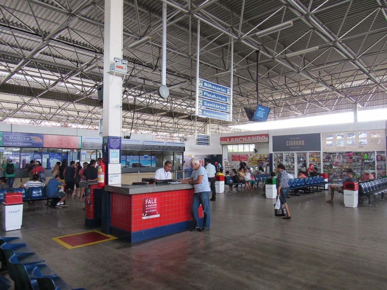 Mercado coberto movimentado com barracas, compradores e um teto metálico alto.