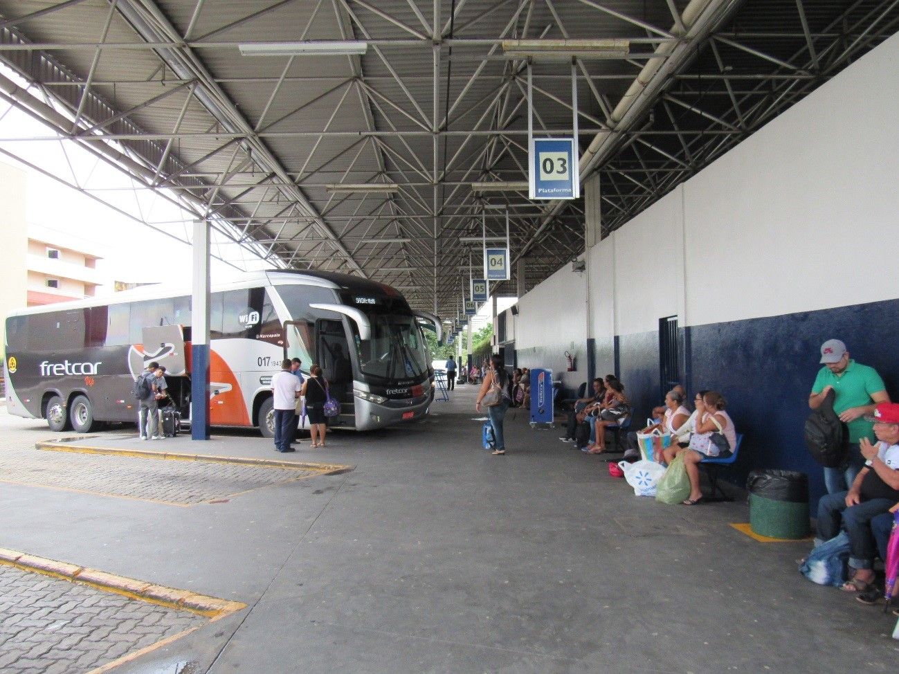 Terminal rodoviário com ônibus estacionados, pessoas esperando sob uma plataforma coberta e bagagens ao lado.
