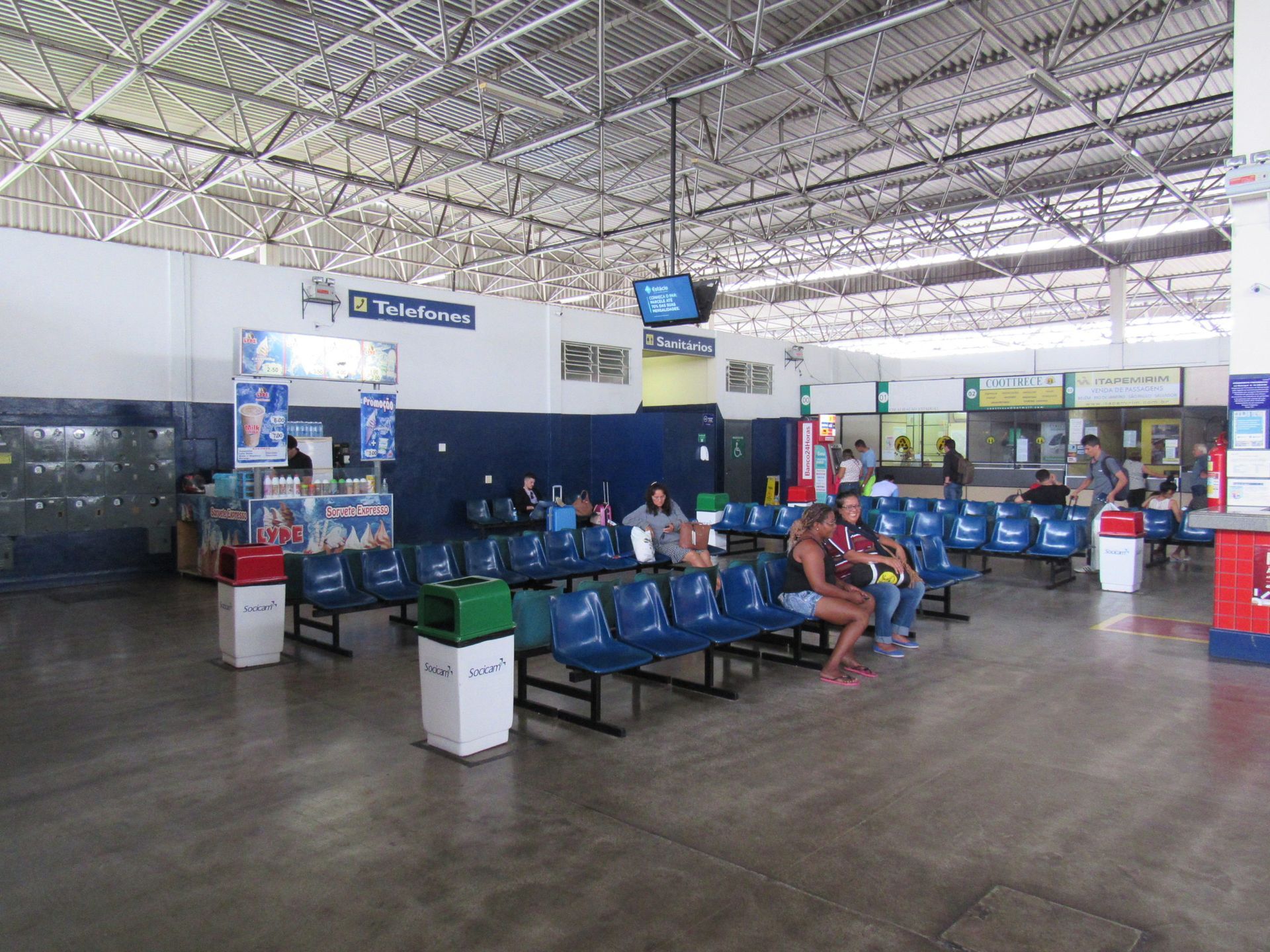 Área de espera vazia do terminal do aeroporto com fileiras de assentos azuis e algumas pessoas sentadas.