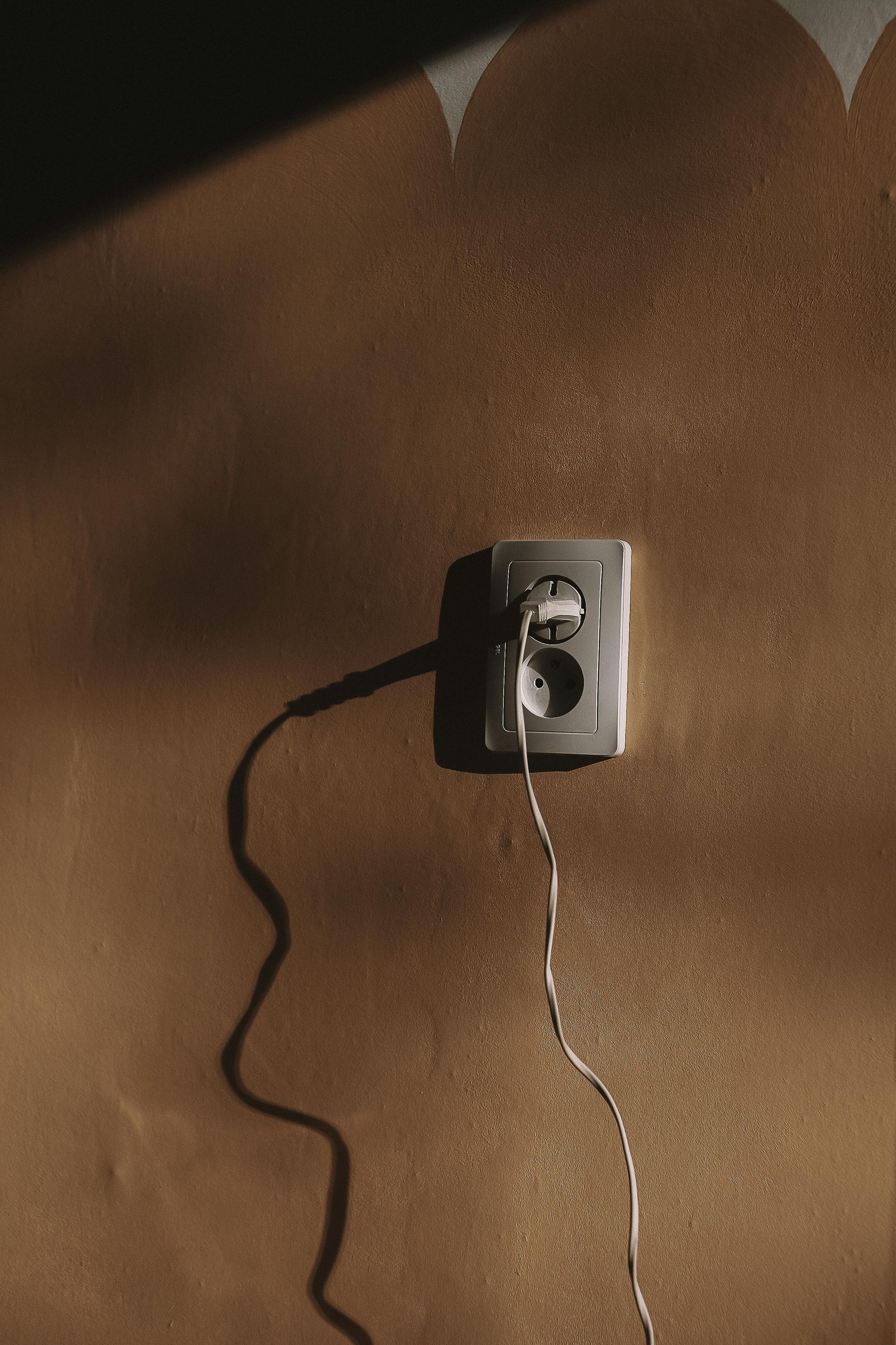 Tomada elétrica com dois fios encostada em uma parede marrom, iluminada por uma sombra forte.