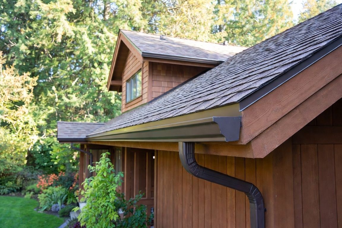 Wooden house roof with dark shingles and a black gutter, surrounded by green trees.