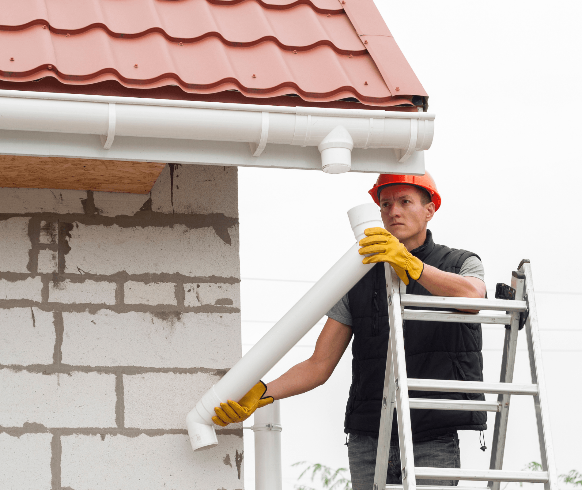 Worker on a ladder installing a white gutter beneath a red roof on a house under construction