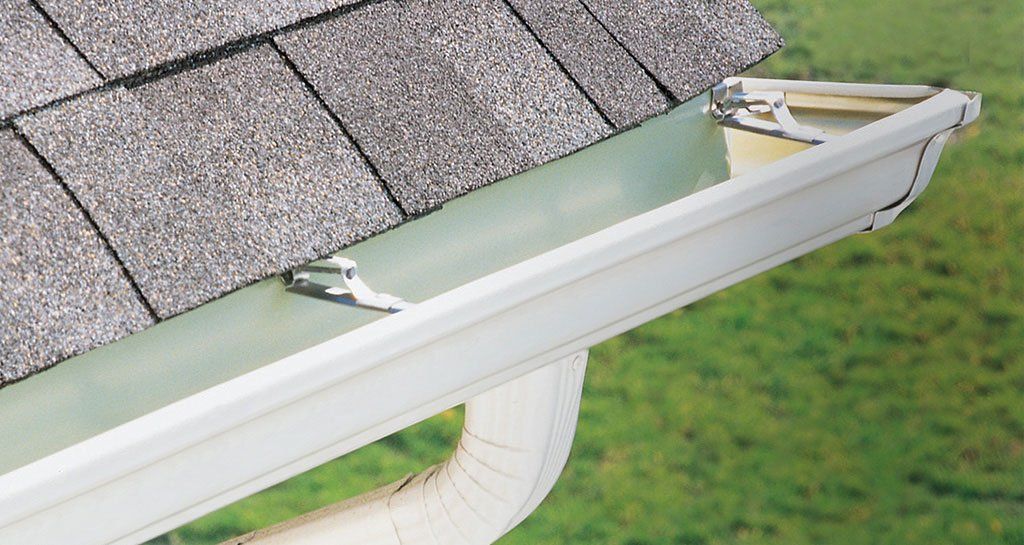 Worker in hard hat and gloves installing white gutter on a house roof beside a ladder.