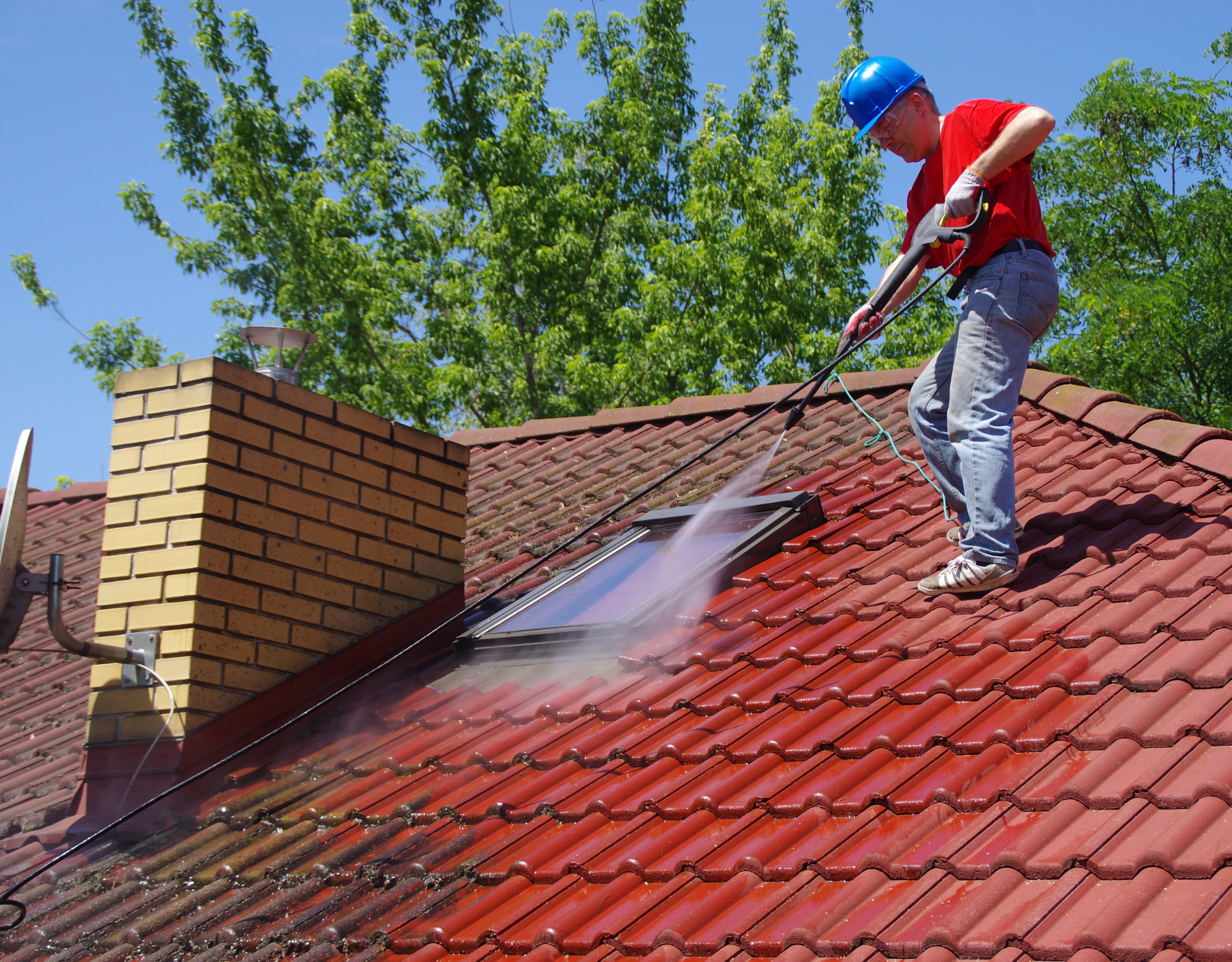 Worker in blue hard hat pressure-washing a red tiled roof beside a brick chimney