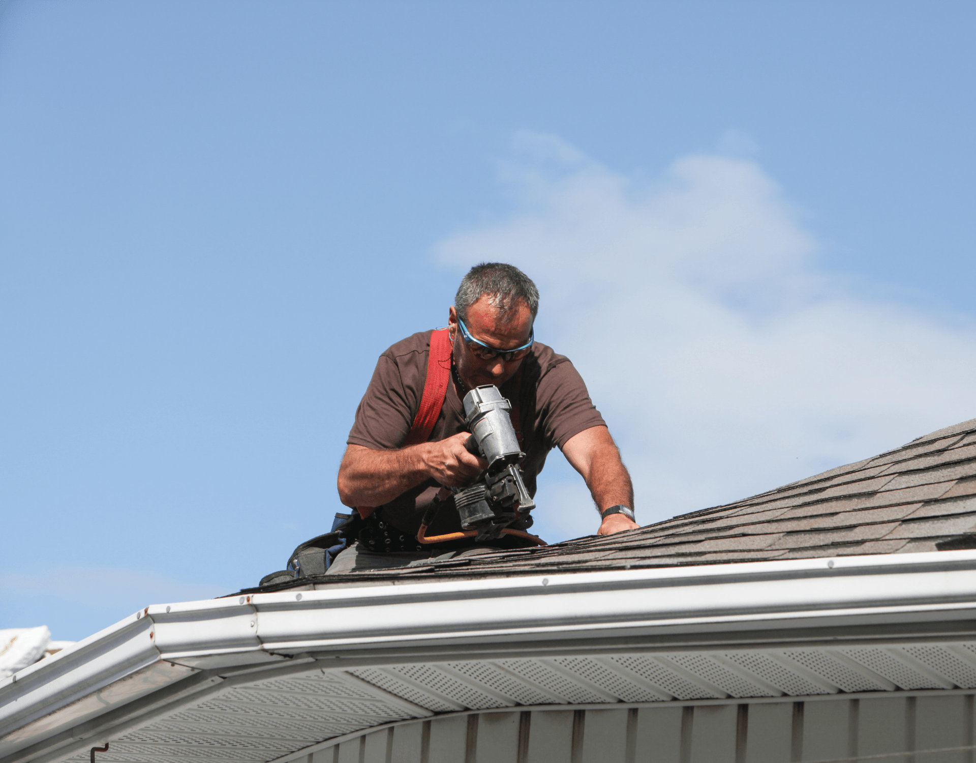 Worker using a nail gun on a shingled roof under a blue sky