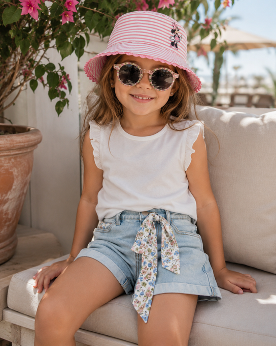 Bambina con cappello da sole rosa e occhiali da sole seduta su un divano all'aperto, sorridente con indosso una maglietta bianca e pantaloncini di jeans.