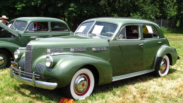 Two vintage green cars parked on grass, one in front with whitewalls and chrome details.