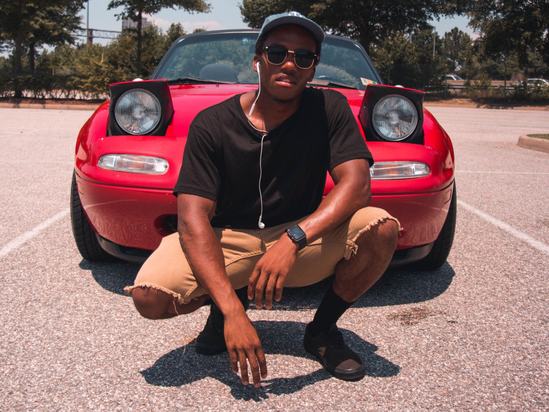 Person crouching in front of a red sports car in a sunny parking lot