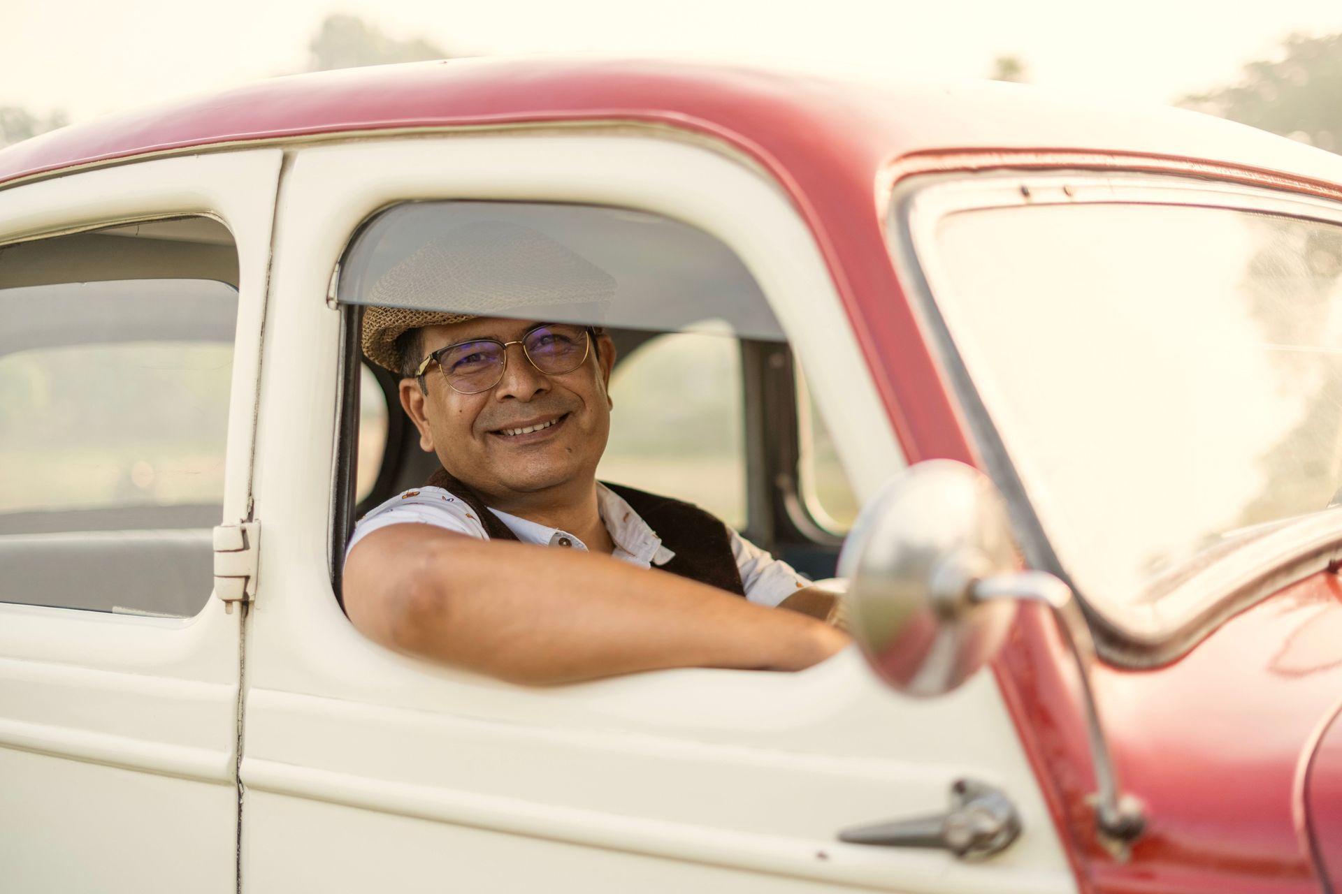 Smiling driver in a red-and-white vintage car, wearing a hat and glasses, looking out the window