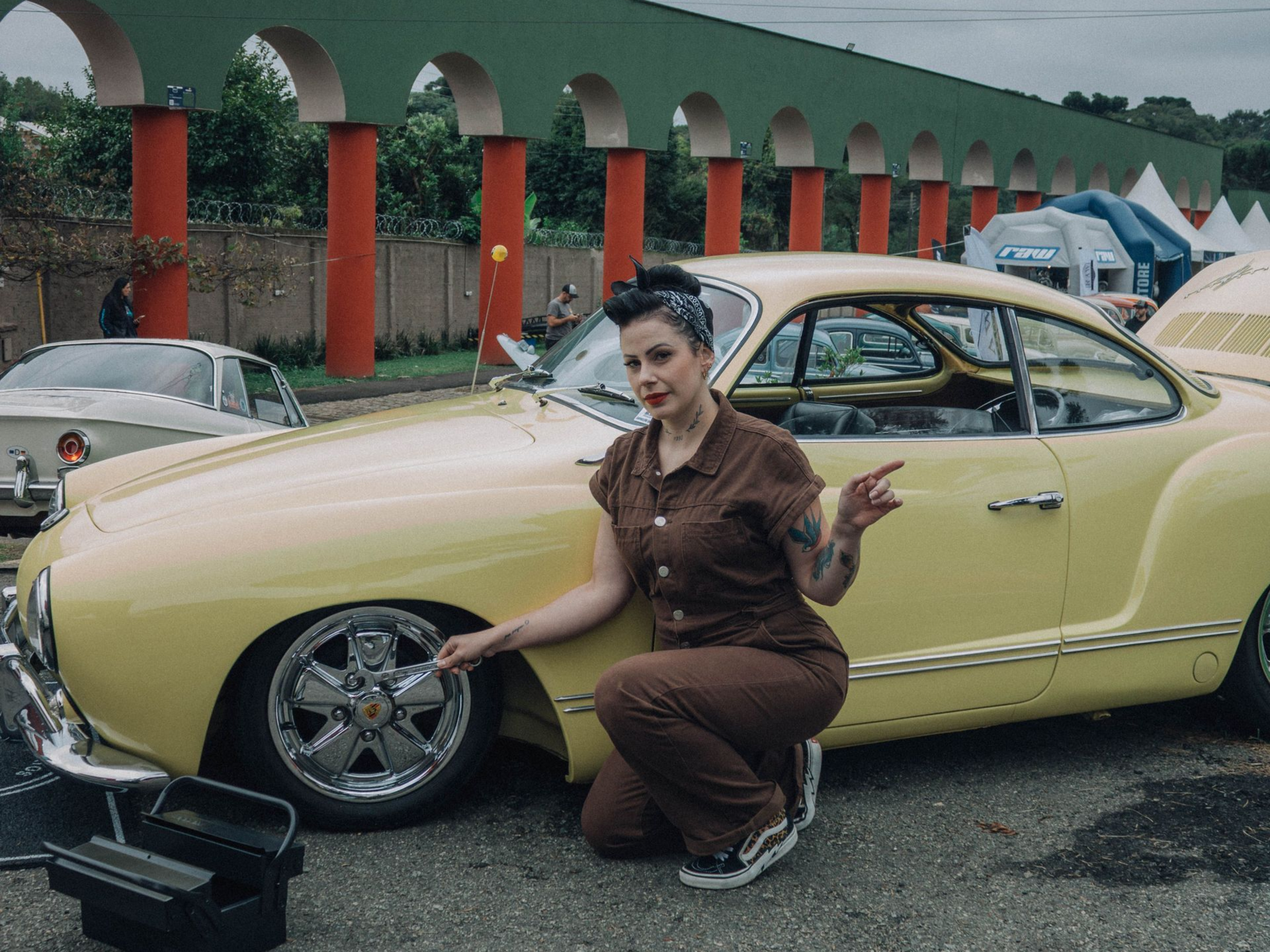 Woman crouches beside a pale yellow classic car at an outdoor car show, gesturing toward its chrome wheel.