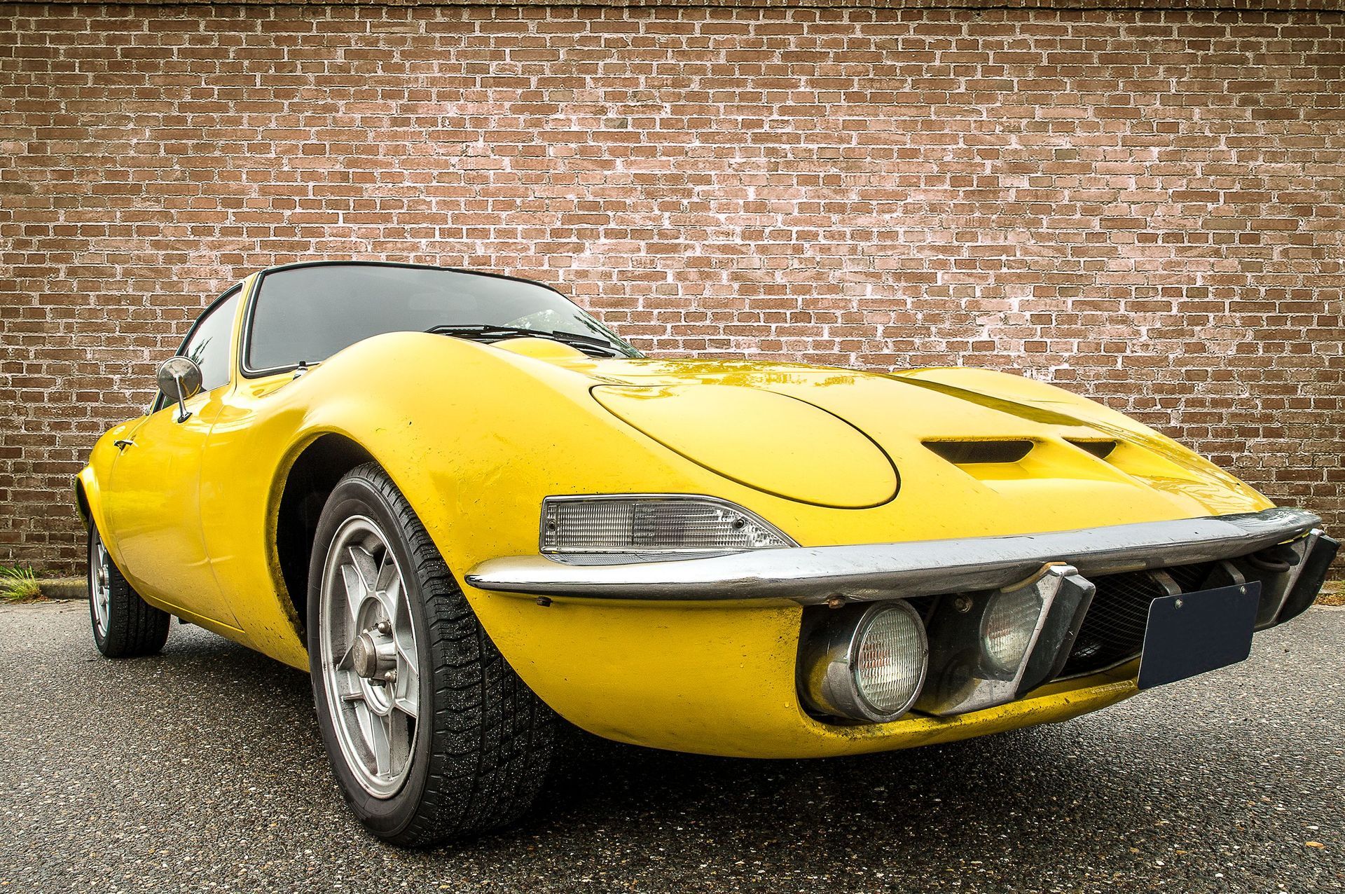 Yellow vintage sports car parked on gravel in front of a brick wall
