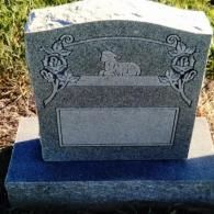 A gray granite headstone featuring an etched image of a resting lamb flanked by floral designs on a blank panel.