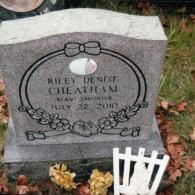 A gray granite headstone for Riley Denist Cheatham, dated July 22, 2010, with floral carvings and a small white fence.