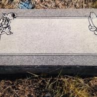 A gray stone grave marker featuring a rose on the left, praying hands on the right, and a blank rectangular space.