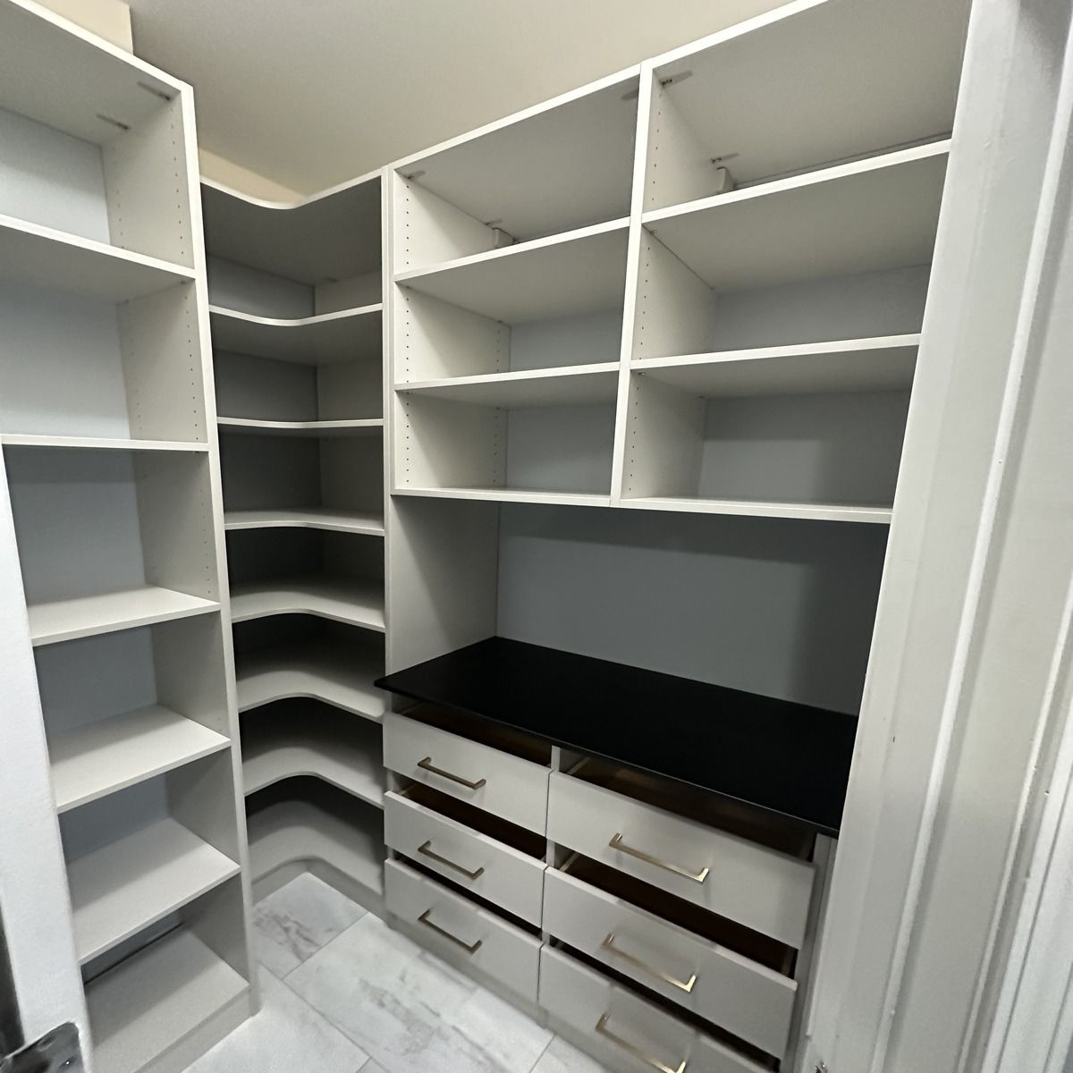 Built-in white closet with shelves, drawers, and a corner section. The drawers have silver handles, and the top has a black surface.