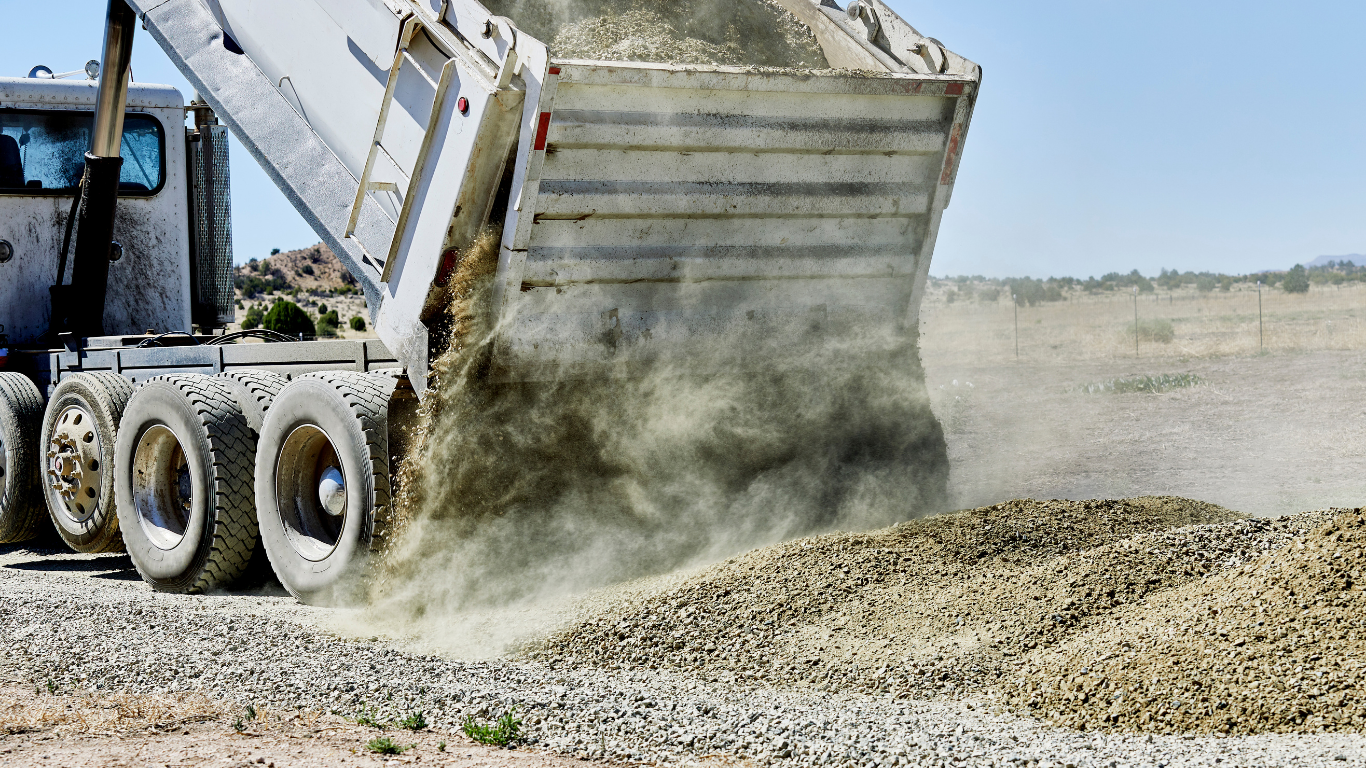 Dump truck unloading gravel on a dirt road, creating dust clouds.
