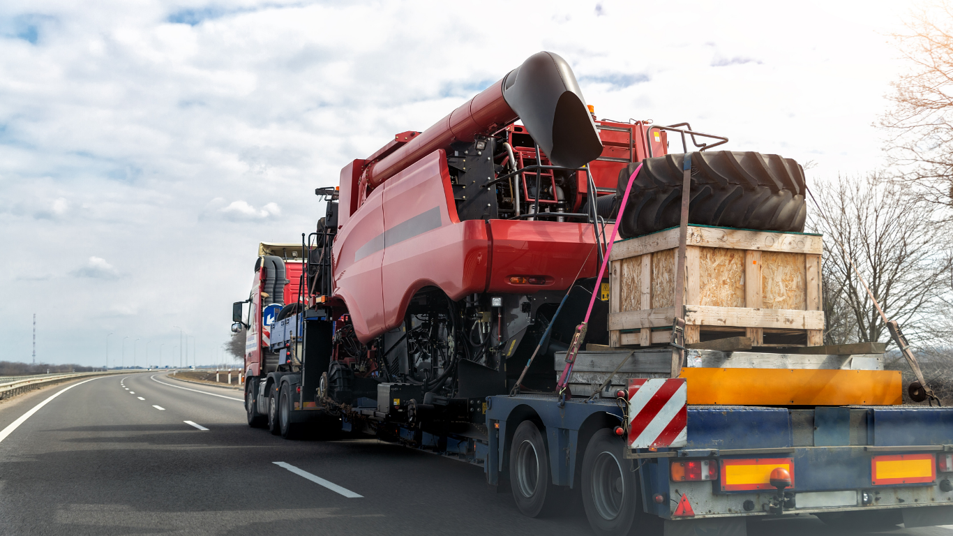 Red combine harvester loaded onto a flatbed truck traveling on a highway under a cloudy sky.