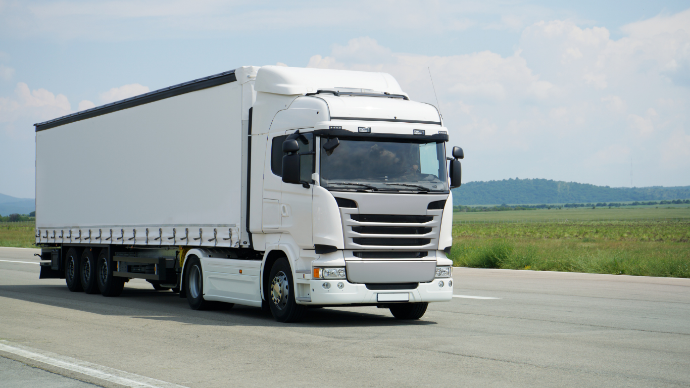 White semi-truck driving on a road with a green field and blue sky in the background.