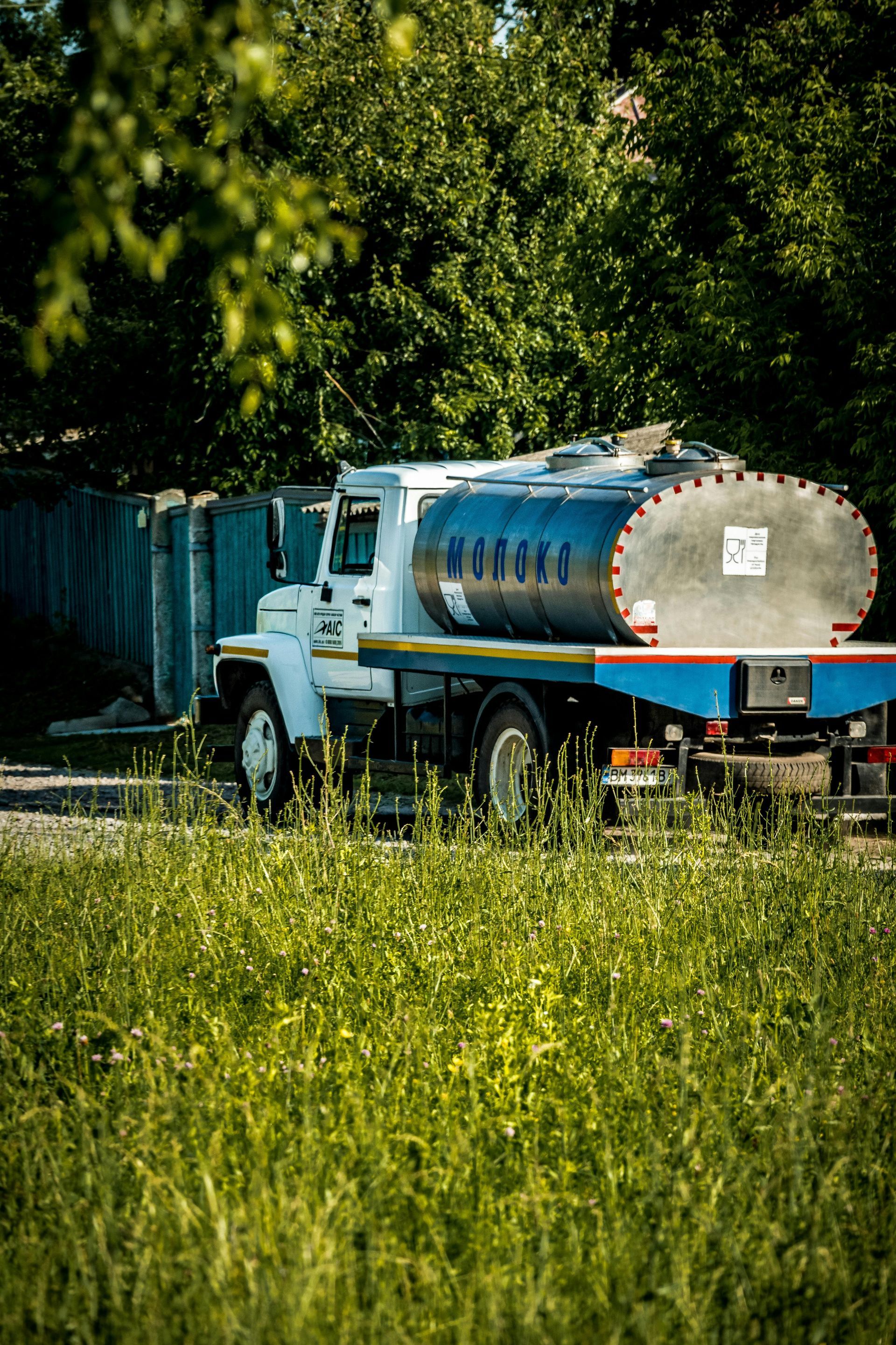 White tanker truck with a silver tank parked on a road, grass and trees in the foreground, wooden fence in background.