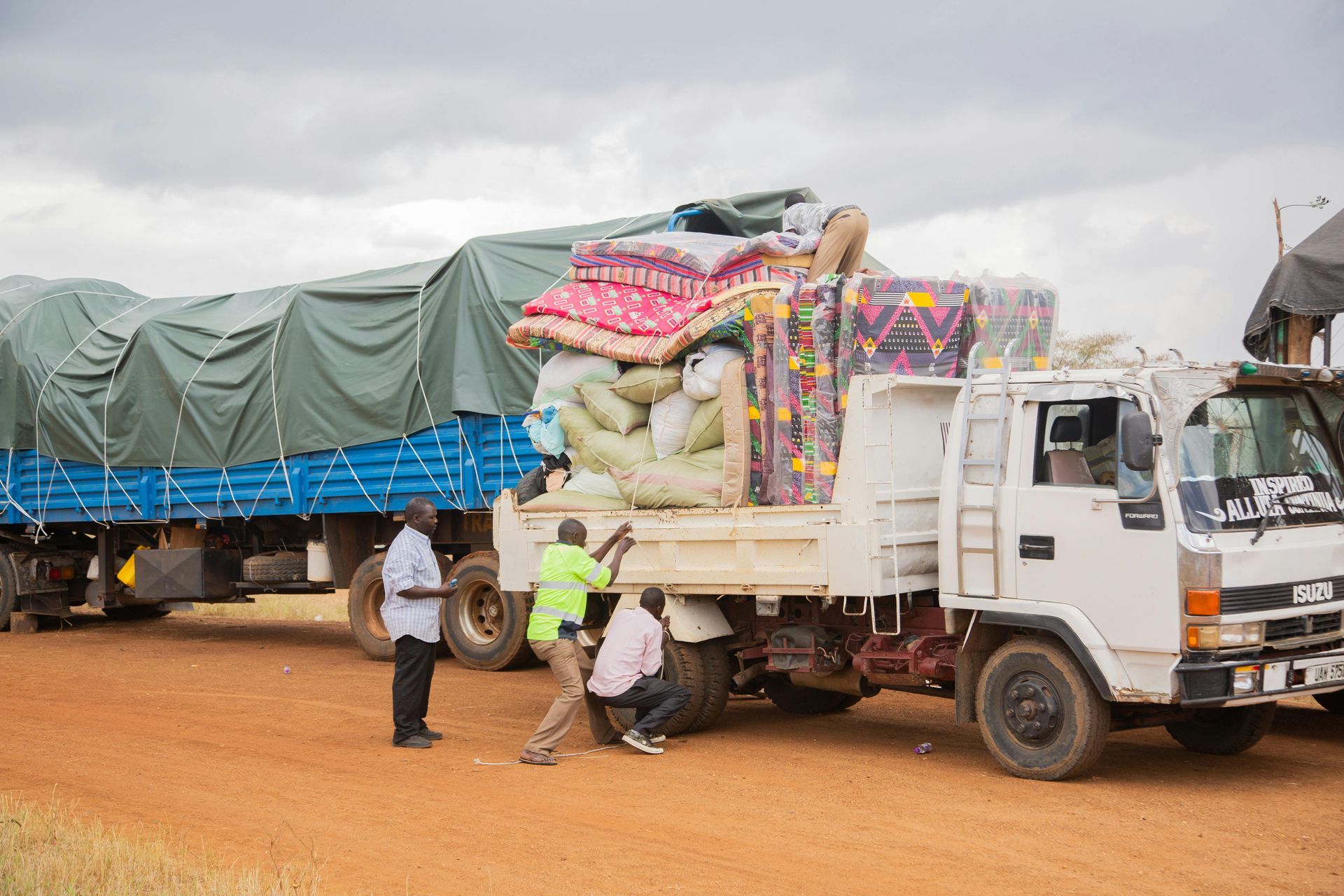 Truck loaded with supplies; men working to secure the cargo. Outdoors, dirt road, overcast sky.