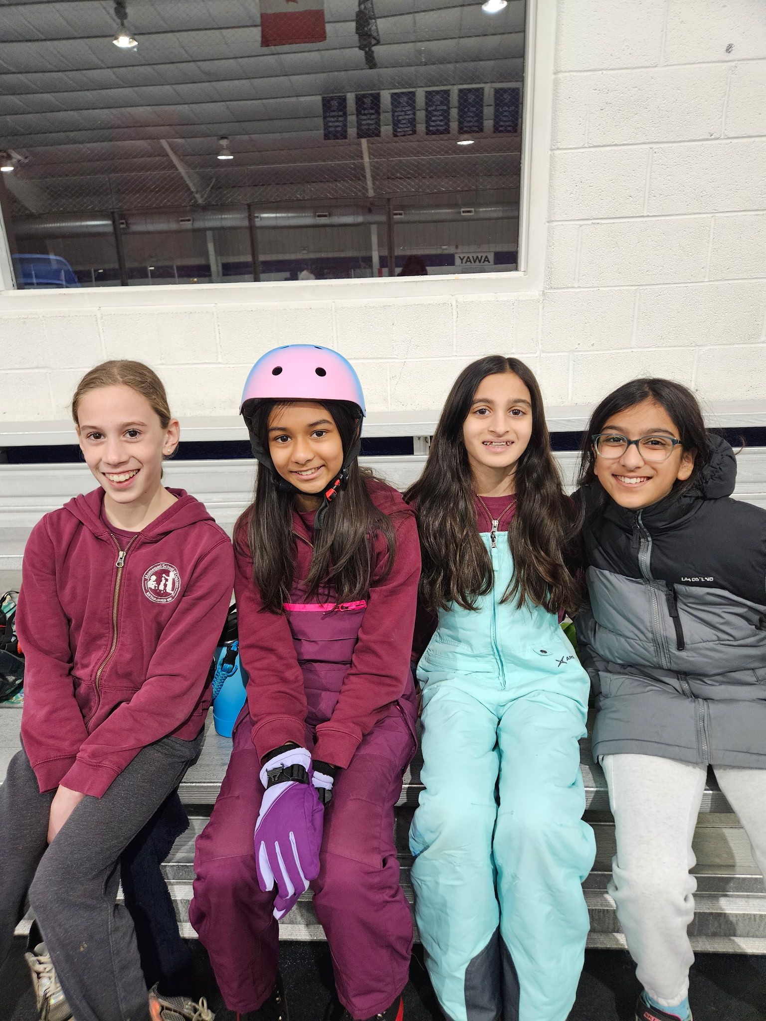 Four friends in winter clothes sit on an indoor ice rink bench, smiling at the camera.