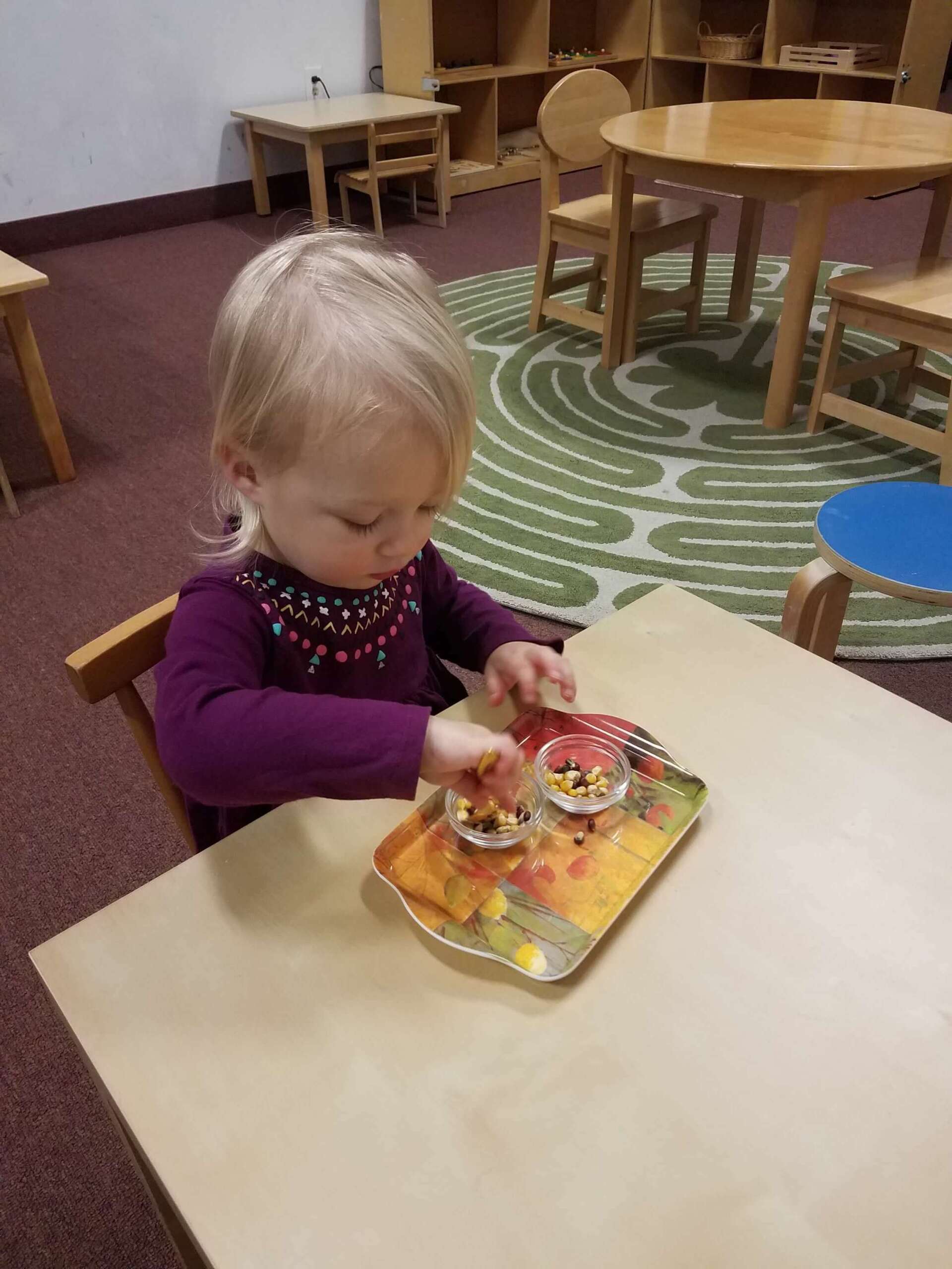 Montessori Young child seated at a table, focused on small cups of snacks on a tray.