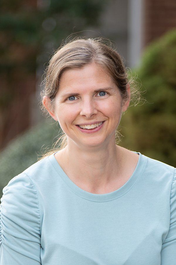 Woman with light brown hair smiles, wearing a light blue top, outdoors.
