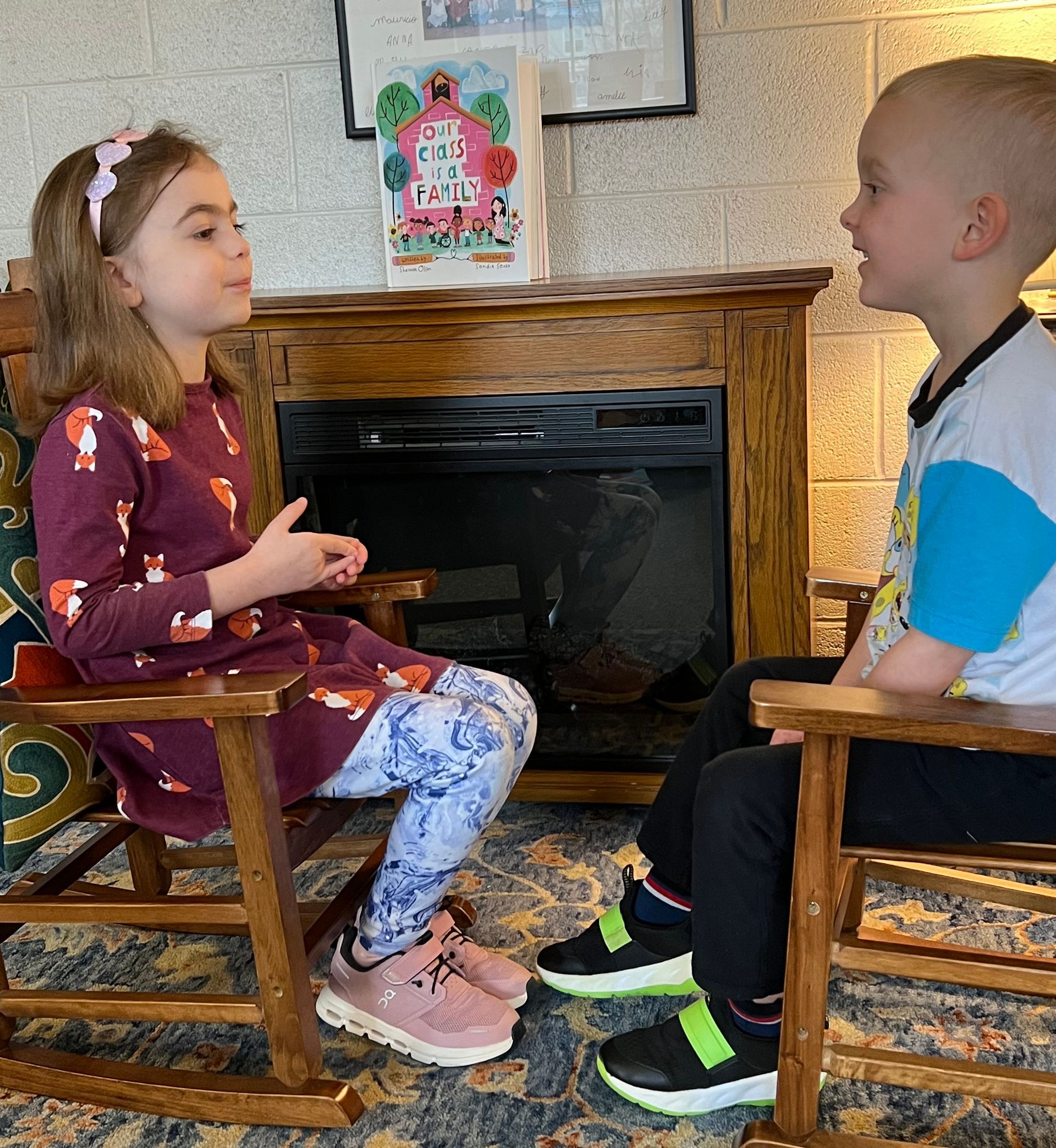 Two children sit facing each other in chairs near a fireplace, talking in a cozy room.