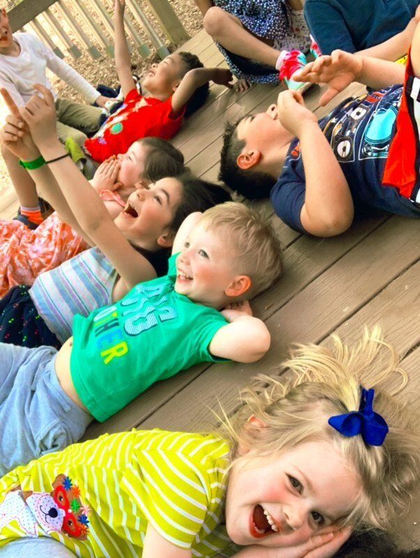 Montessori children lying on wooden deck, looking and pointing upwards with happy expressions.
