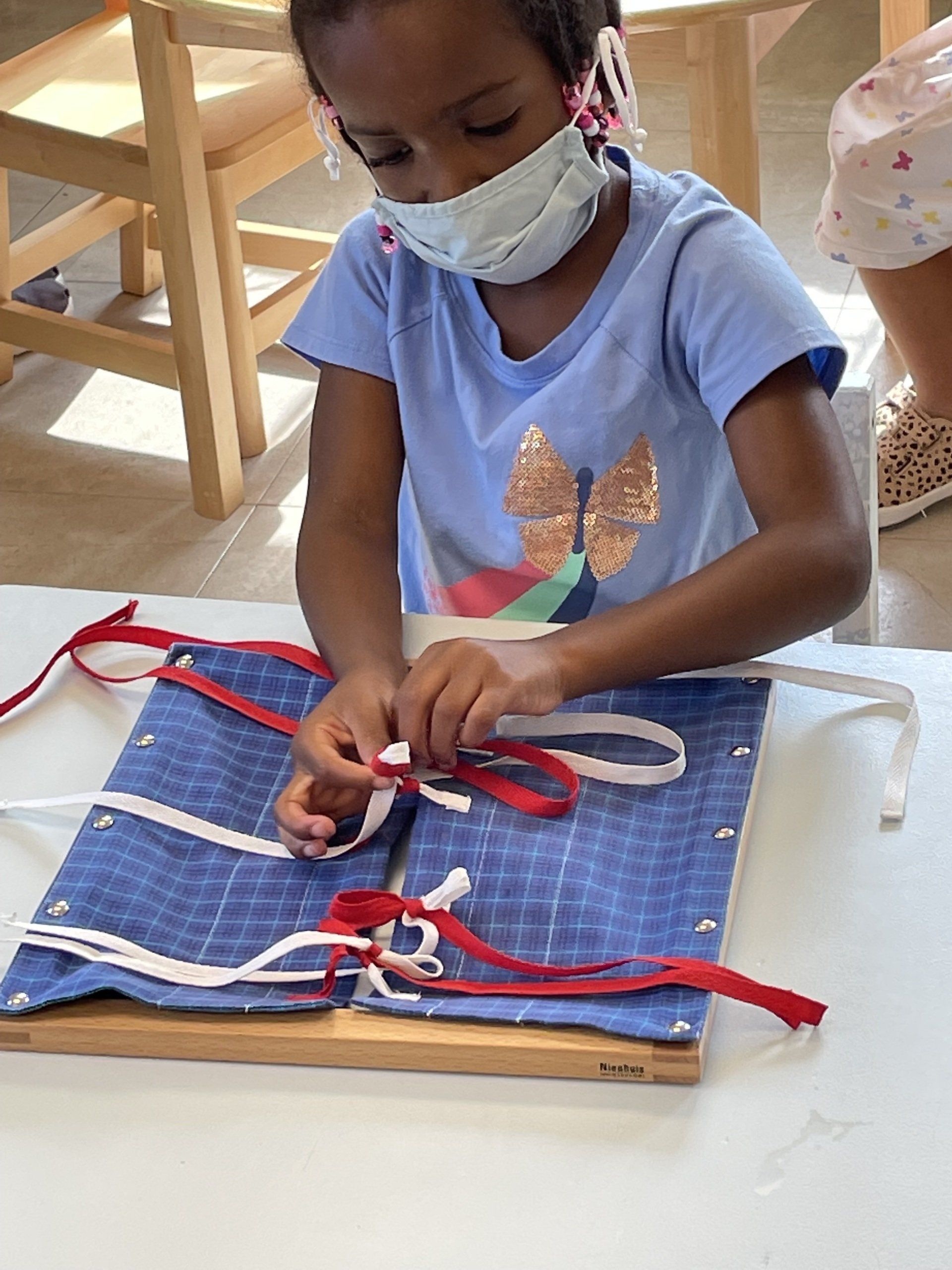 A Montessori child wearing a mask is tying ribbons on a wooden board.