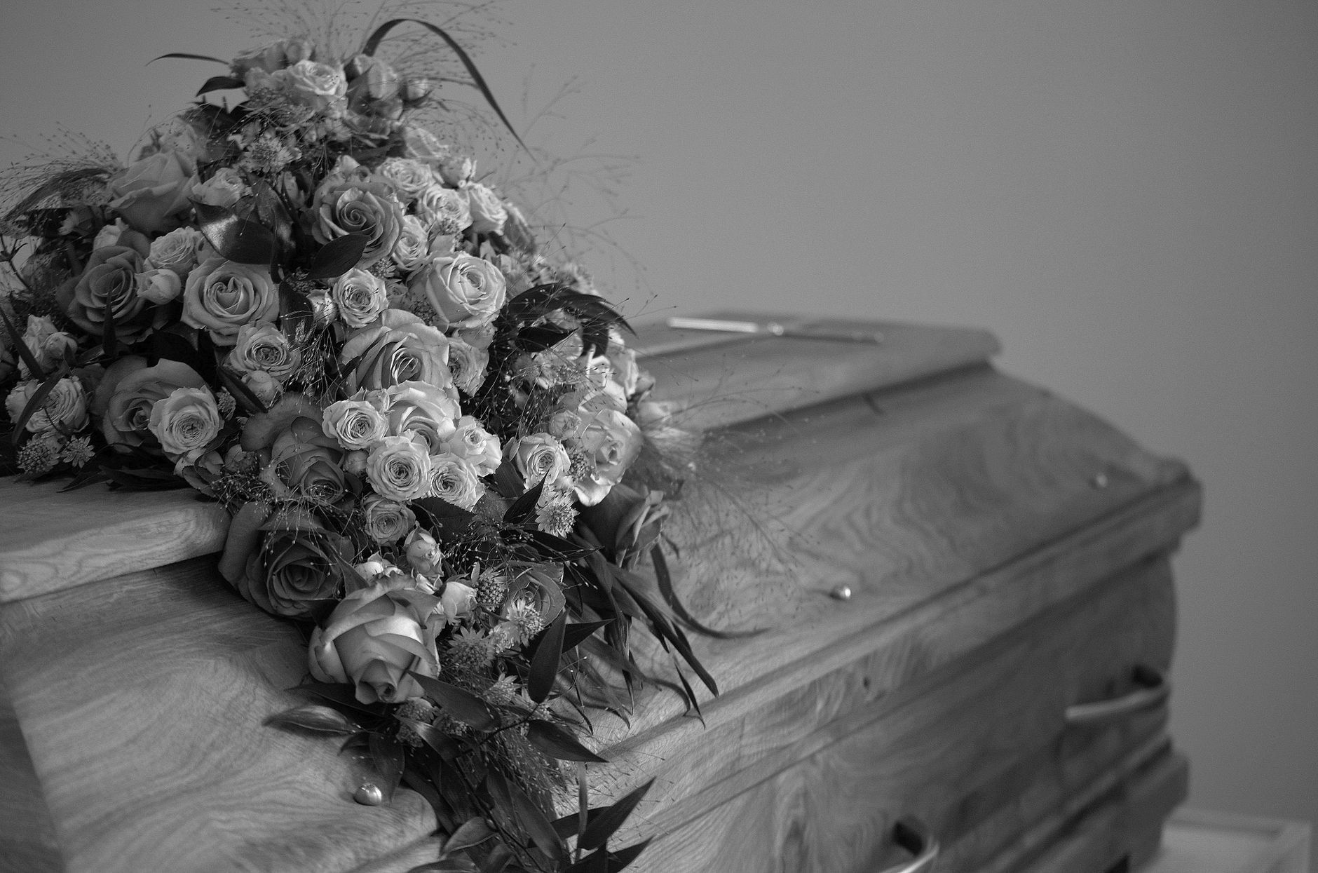 A black-and-white photograph of a wooden casket draped with a large rose bouquet, set against a plain wall in dim, somber interior light.