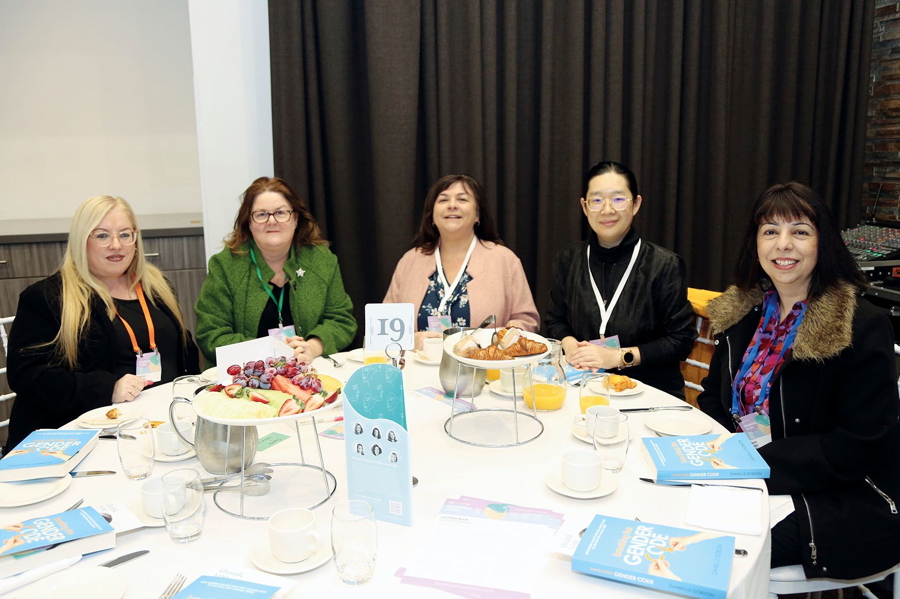 Five women seated at a round table, possibly at an event. They smile at the camera, with food and drinks on the table.