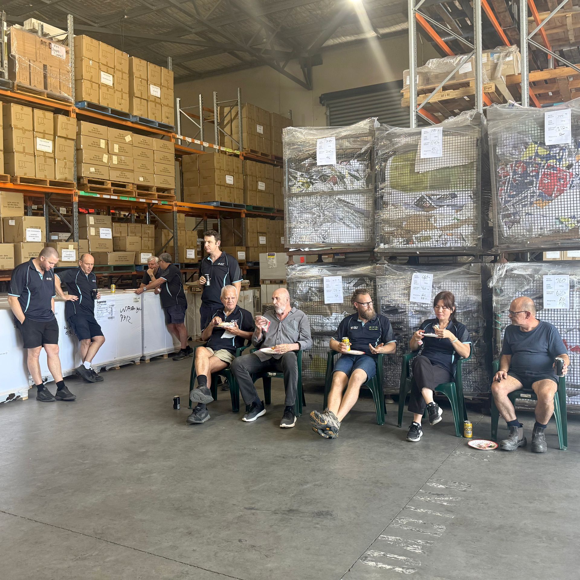 A group of people in a warehouse, some sitting and eating, others standing near stacks of boxes and pallets.