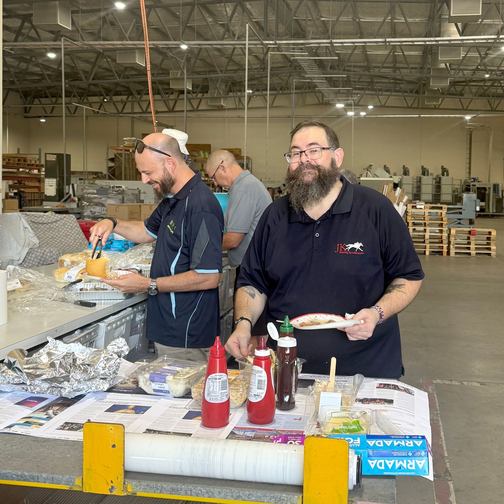 Men serving and eating hot dogs at a casual gathering in a warehouse. Ketchup and mustard bottles are visible.
