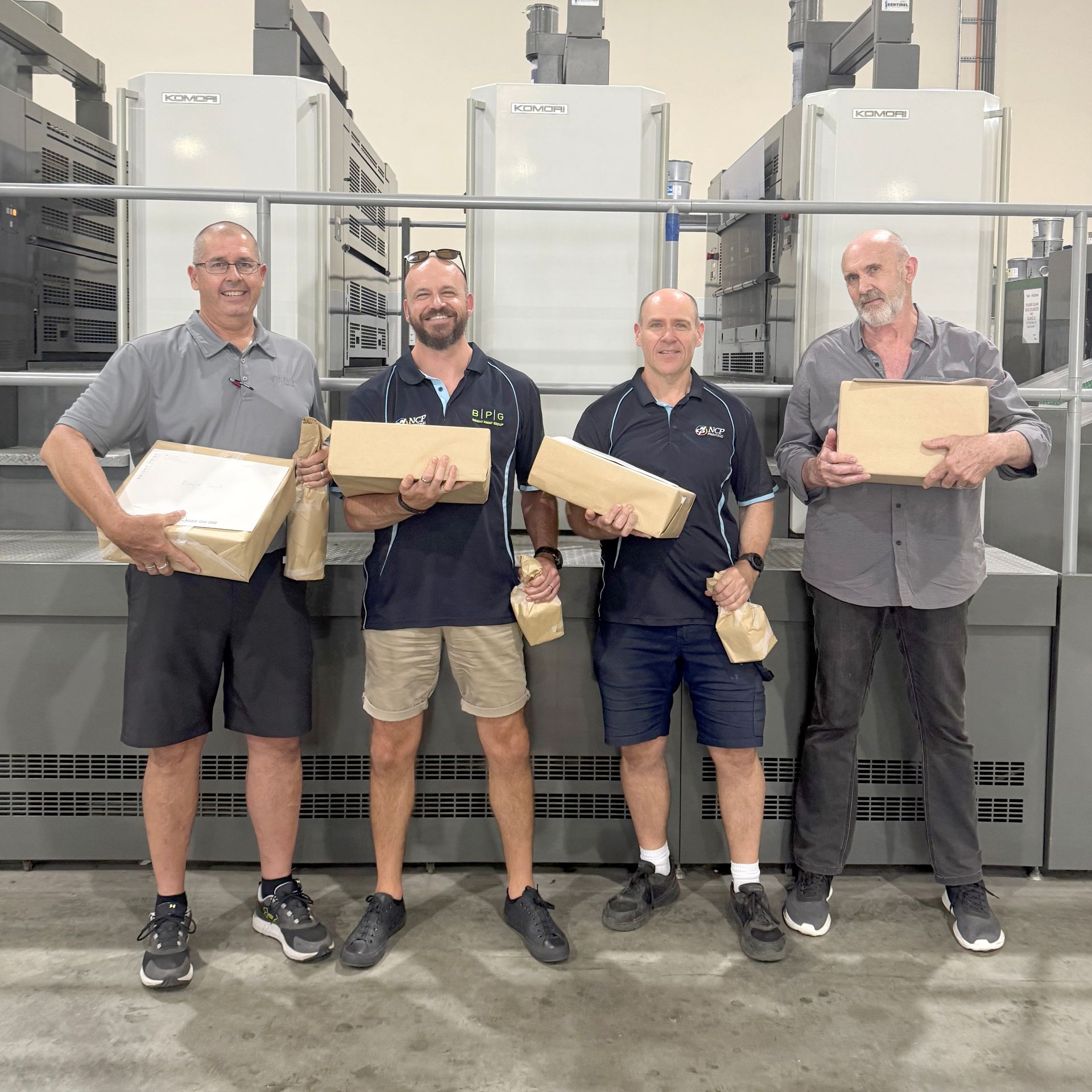 Four men holding boxes and packages stand in front of large printing machines in a factory setting.
