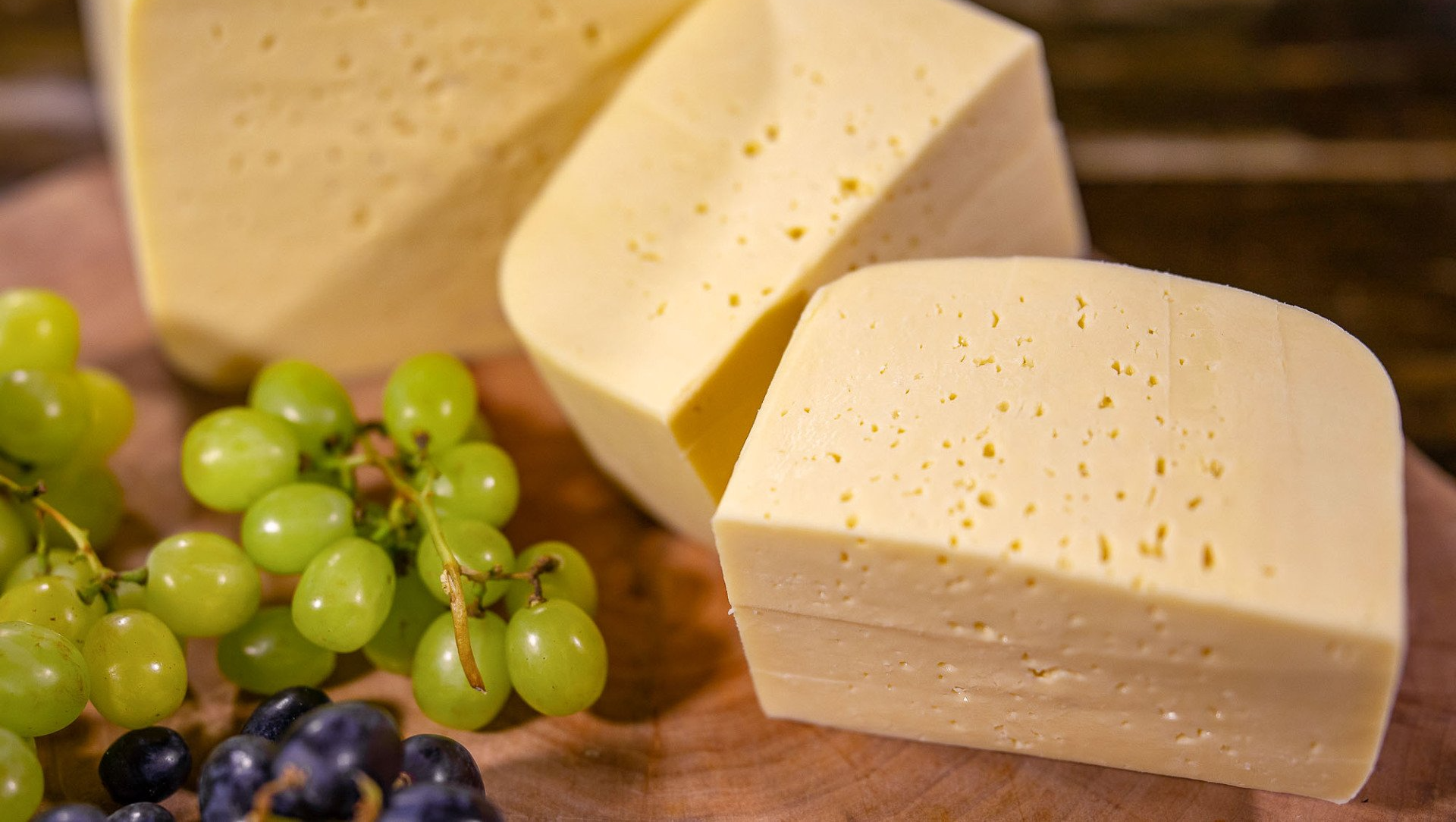 Drei Stücke Hofkäse mild liegen neben Weintrauben auf einem Holzschneidebrett.