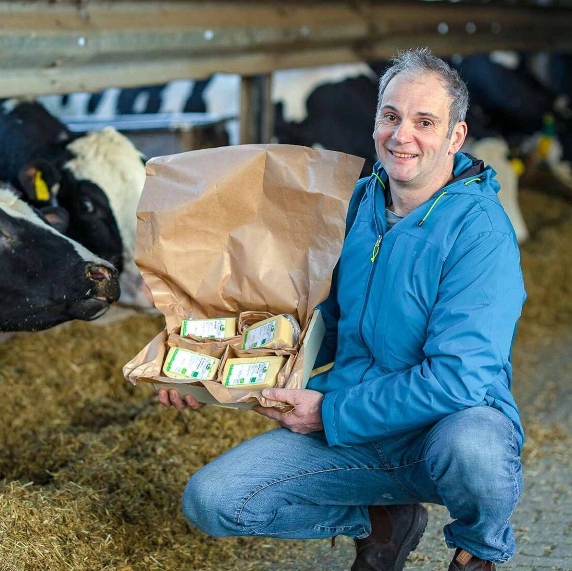 Ludger Engeln in blauer Jacke kniet vor Kühen im Stall und hält einen Vesandkarton mit 4 Stücken Engelnhof Käse in der Hand.