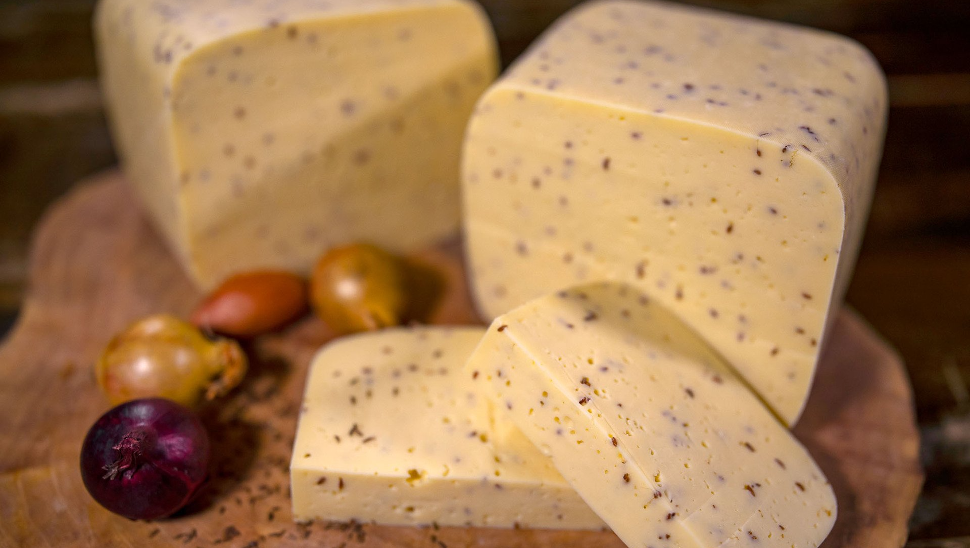 Mehrere Stücke Käse mit Kümmel liegen neben Zwiebeln auf einem Holzschneidebrett.