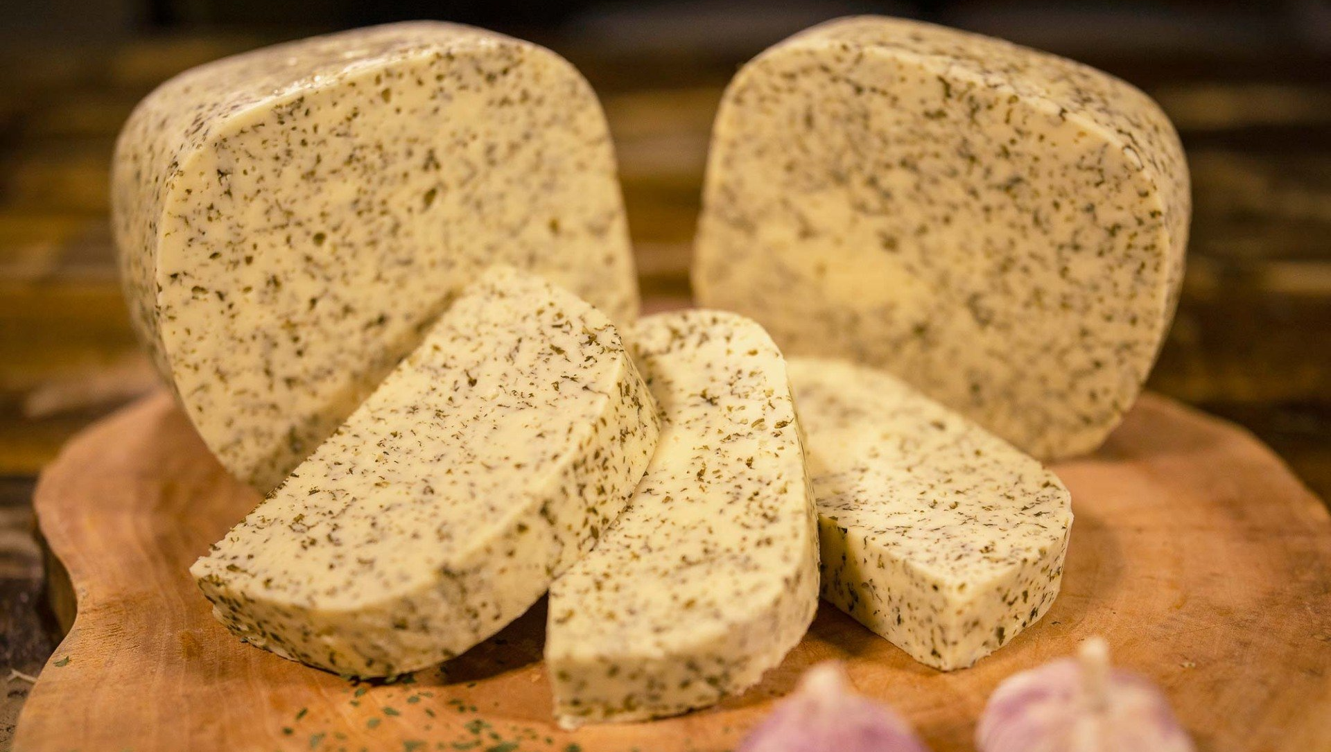 Mehrere Stücke Hofkäse mit Bärlauch liegen auf einem hölzernen Schneidebrett. Davor  zwei Knoblauchknollen.