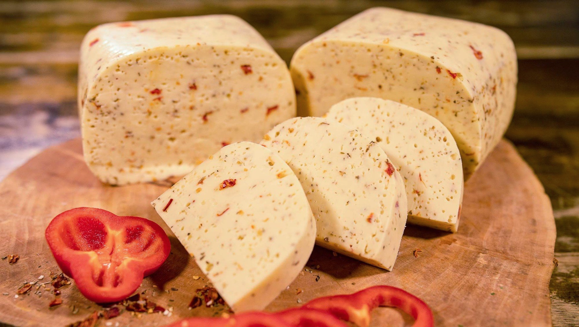 Mehrere Stücke Hofkäse mit italienischen Kräutern liegen auf einem hölzernen Schneidebrett. Davor liegt geschnittene Paprika.