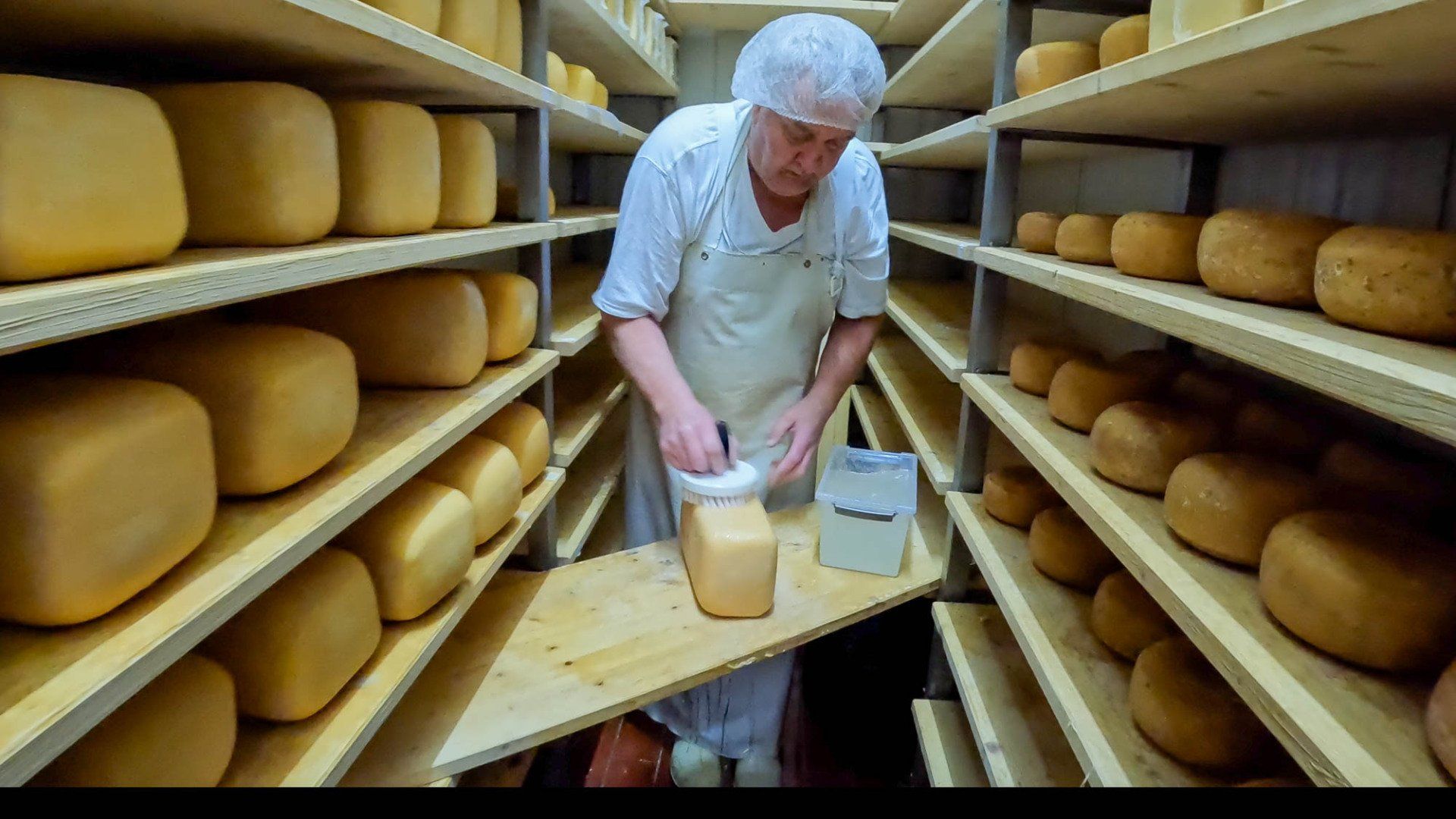 Ein Mann in einer weißen Schürze steht in einem Raum voller Käseregale und bestreicht einen Laib Käse auf einem Holzbrett.