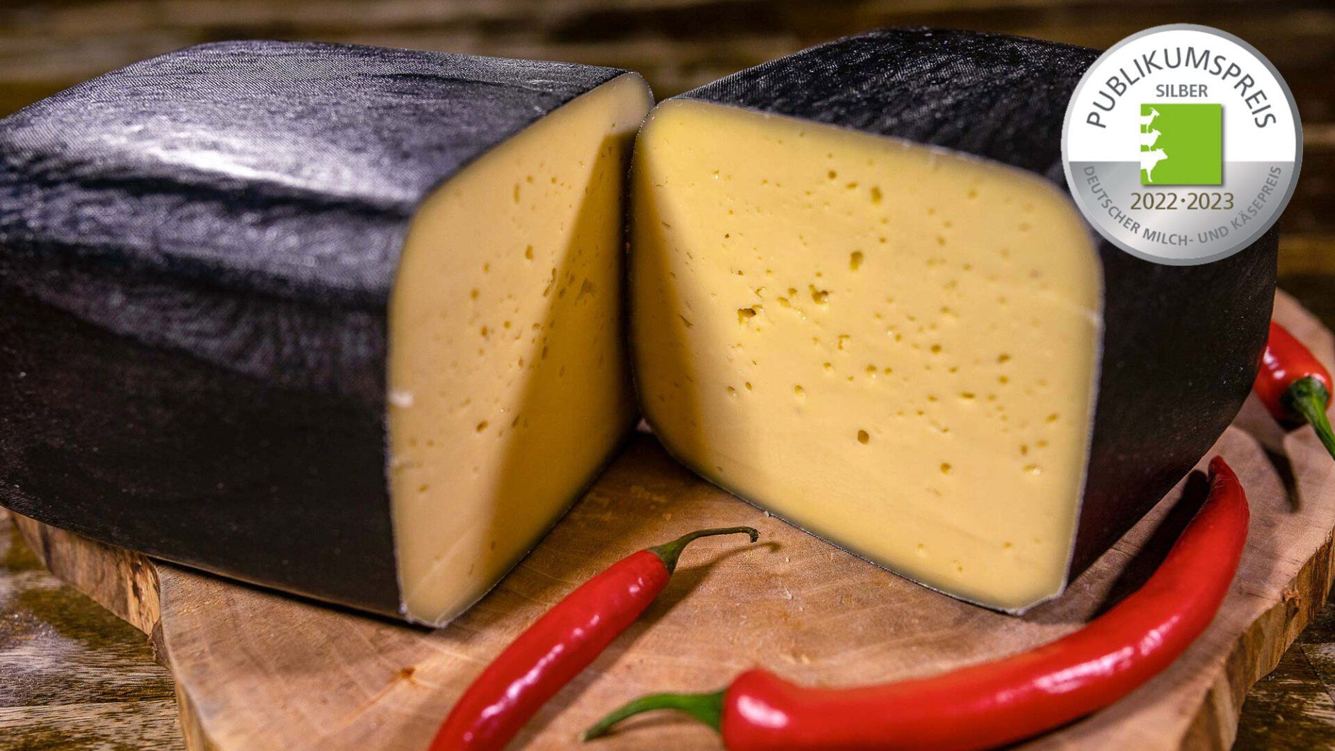 Ein Laib Moorkäse liegt angeschnitten auf einem Holzschneidebrett neben roten Peperoni.