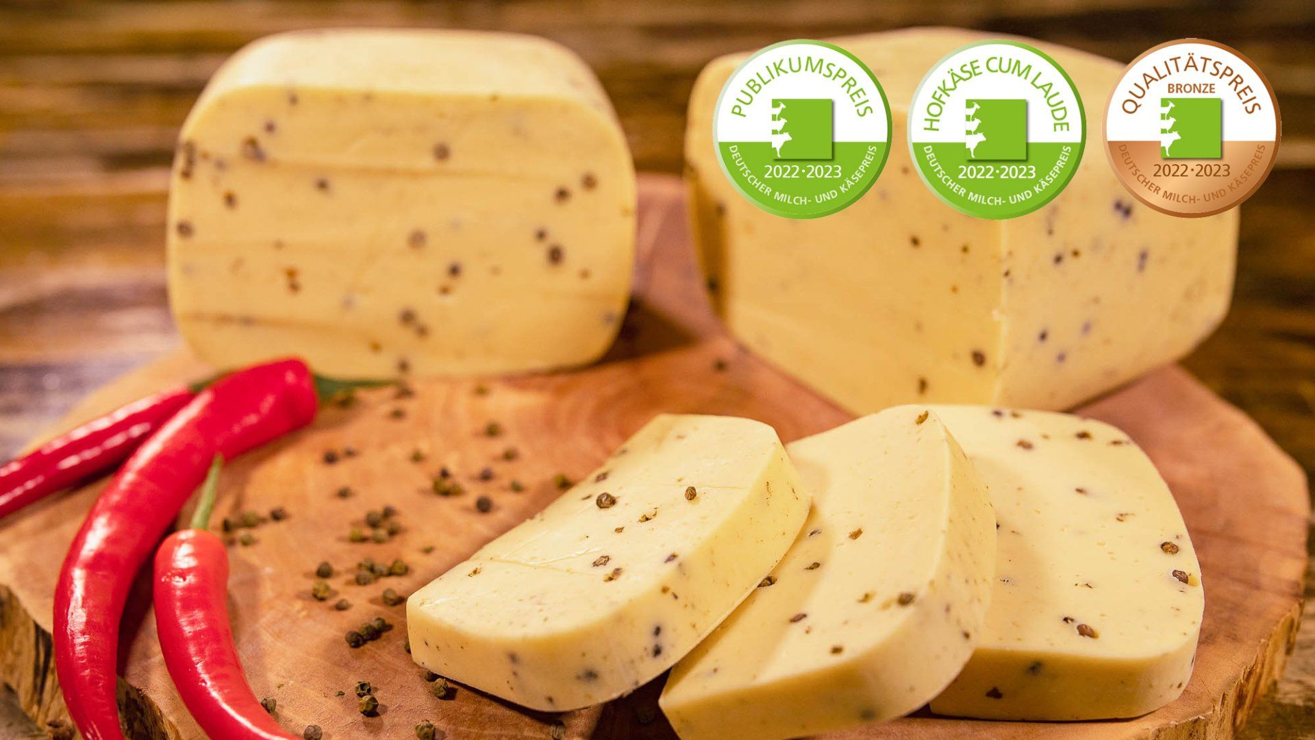 Ein hölzernes Schneidebrett mit 5 Stück Hofkäse mit Pfeffer. Daneben Pfefferkörner und rote Peperoni. 