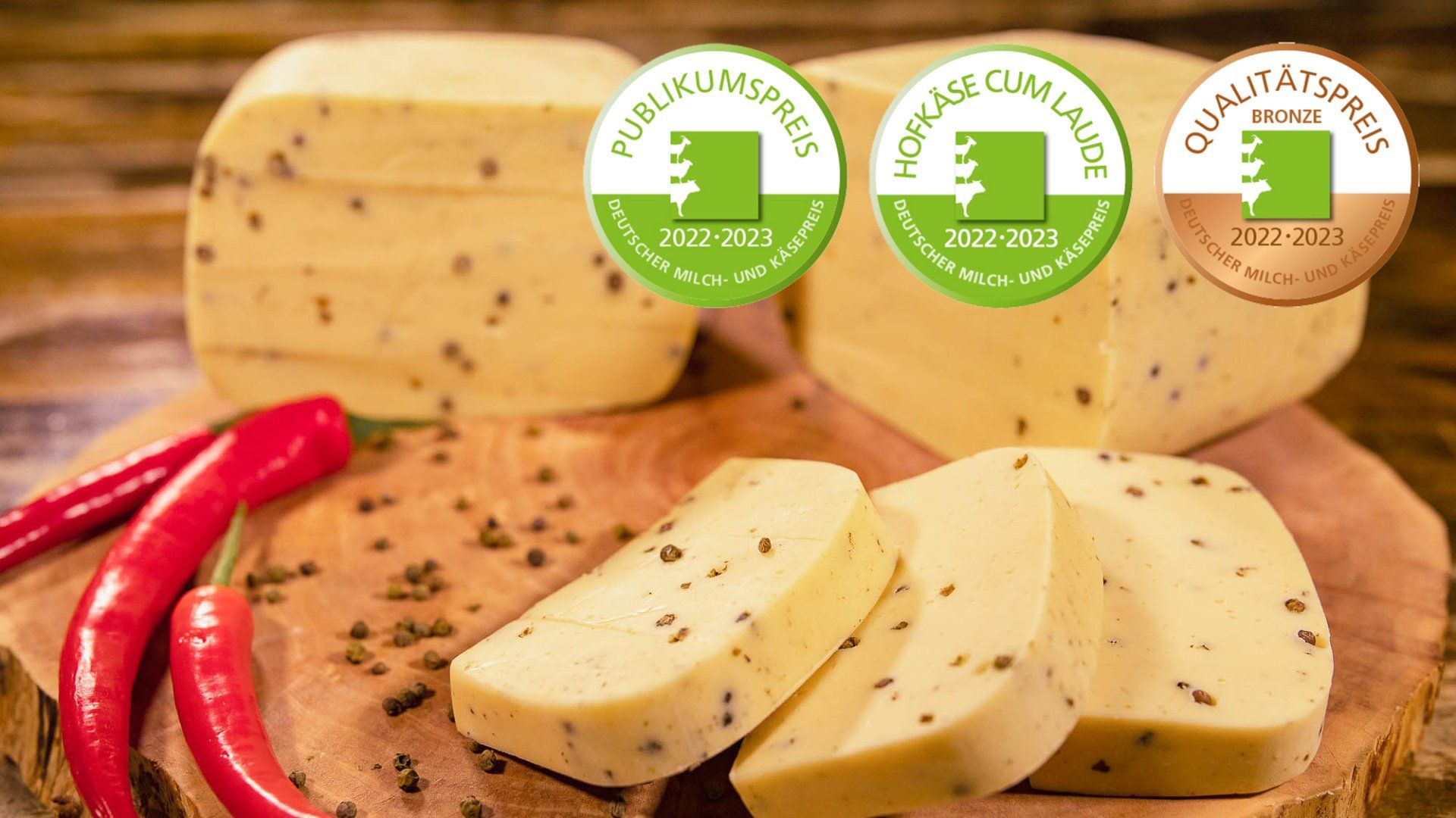 Ein hölzernes Schneidebrett mit 5 Stück Hofkäse mit Pfeffer. Daneben Pfefferkörner und rote Peperoni. 
