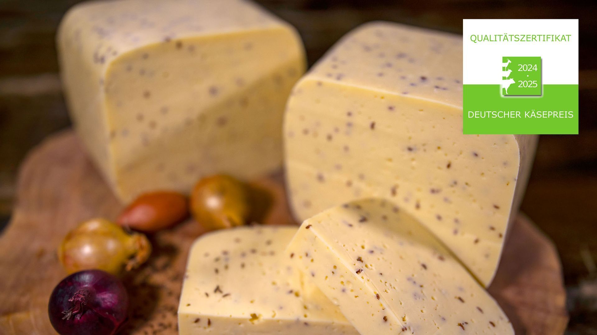 Mehrere Stücke Käse mit Kümmel liegen neben Zwiebeln auf einem Holzschneidebrett.