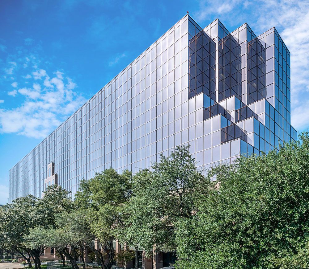 Modern glass office building with reflective windows and trees in front under a blue sky