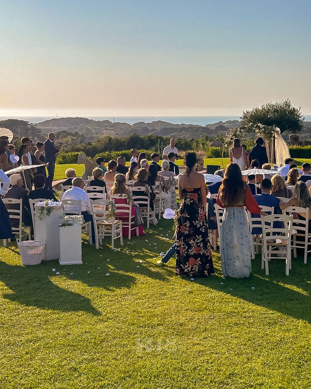 Gli invitati siedono su sedie bianche a un matrimonio all'aperto al tramonto, su un prato con vista sulla città.