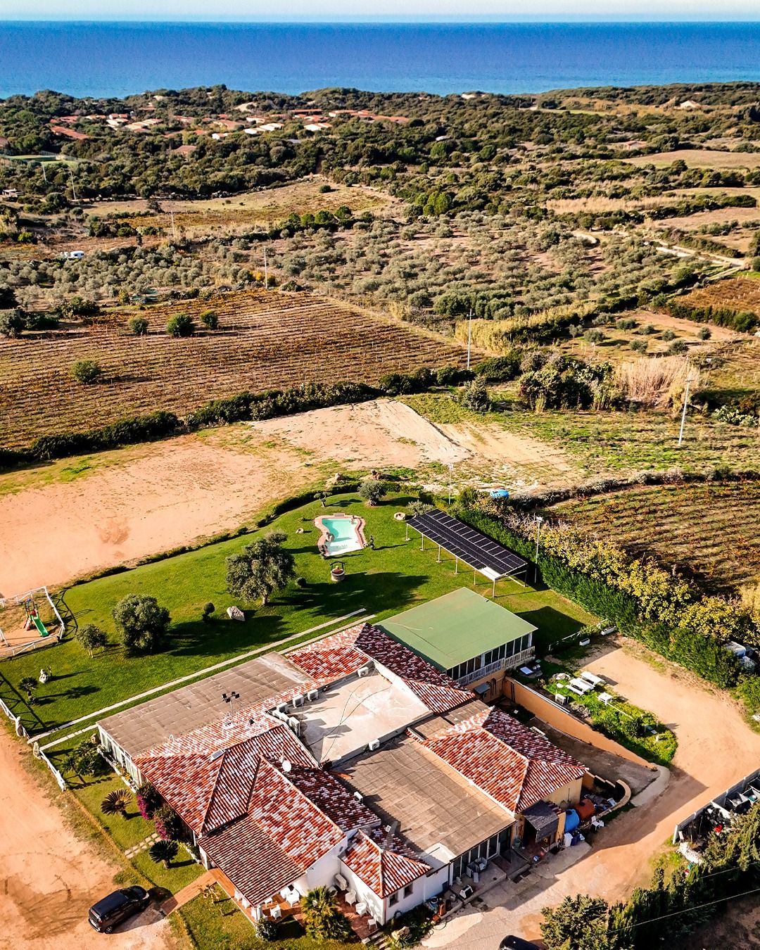 Veduta aerea di una casa con tetto di tegole rosse in un paesaggio rurale costiero, con campi e l'oceano sullo sfondo.