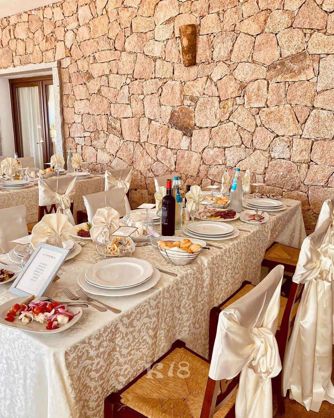Elegante tavolo da banchetto apparecchiato con tovaglie bianche, piatti e bottiglie, addossato a un muro di pietra.