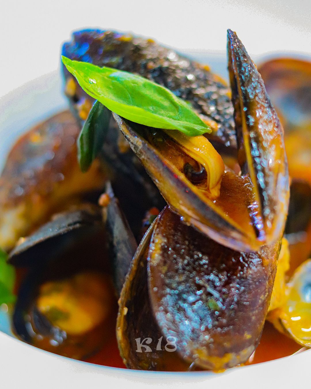 Primo piano di cozze al vapore in una ricca salsa marrone, guarnite con una foglia di erba aromatica verde.